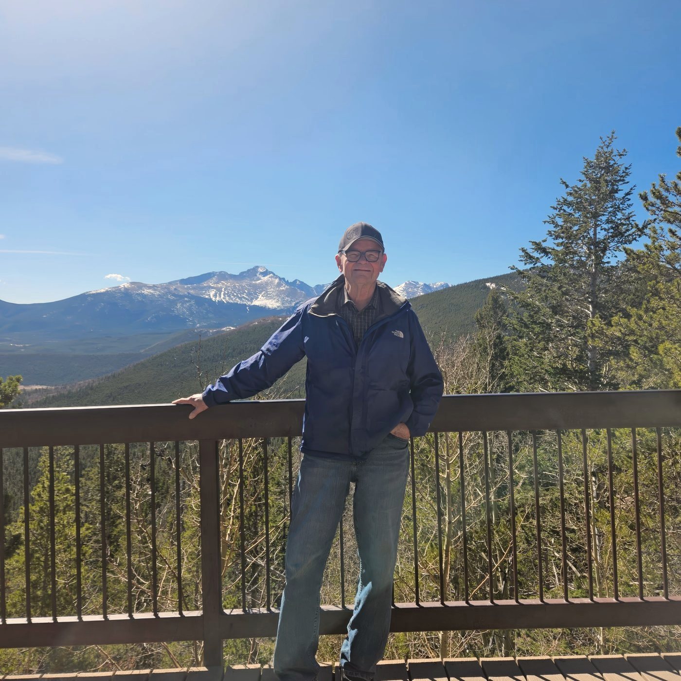 Bob King standing at a railing overlook with snow-capped Rocky Mountains behind him