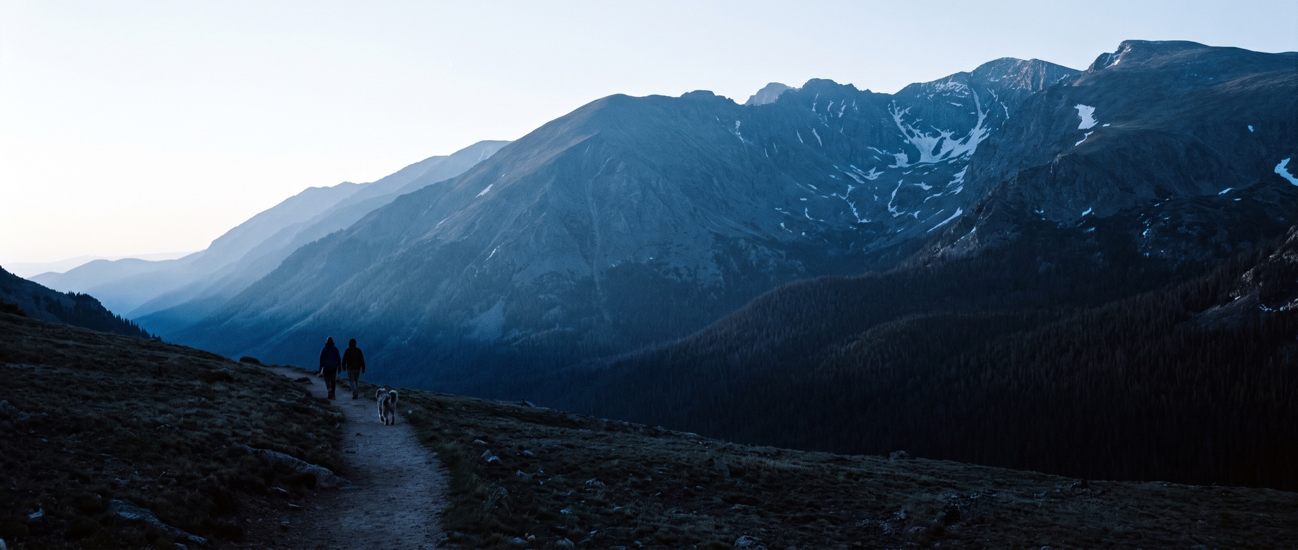 Two distant figures and a dog walking a mountain trail at dawn, layered Colorado ridgelines ahead of them
